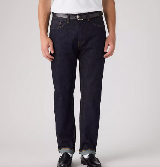 A person wearing dark indigo straight-leg jeans with a black belt, styled with a white shirt and black leather shoes, photographed against a light gray studio background.