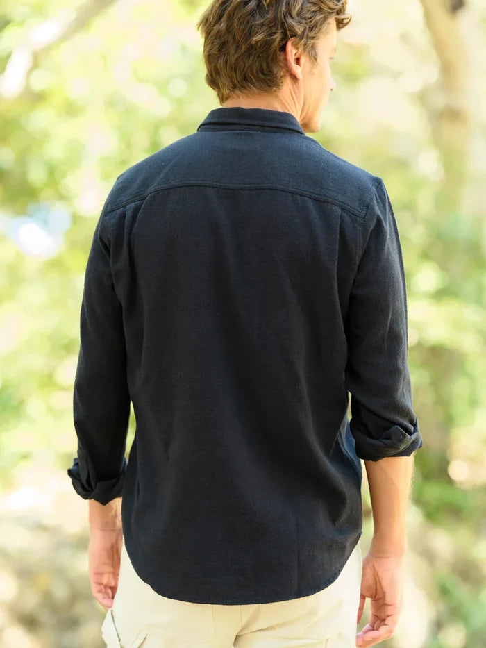 Model wearing Mollusk Surf Shop One Pocket Shirt in Black Flannel, back view walking in dappled outdoor light showing deep black brushed 95% cotton 5% linen flannel, point collar, clean back yoke, long sleeves rolled to mid-forearm, and slight saddle curve hem, styled with cream trousers