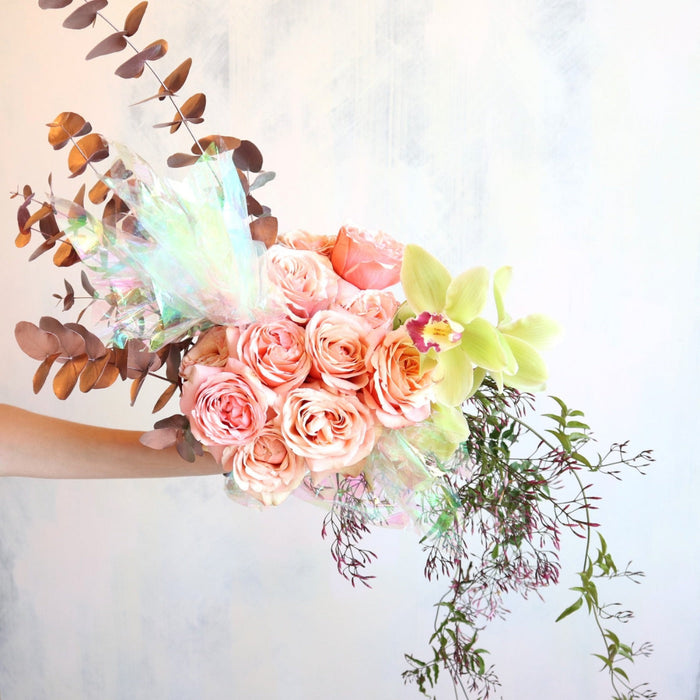 A bouquet of pink roses with green and brown foliage, wrapped in an orchid, with iridescent accents.